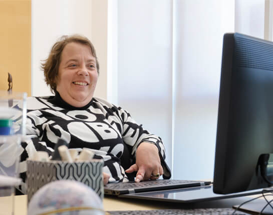 Helle Deertz sitzt am Schreibtisch. Sie trägt einen schwarz-weißen Pullover und lächelt in die Kamera. Im Vordergrund ein Computerbildschirm.