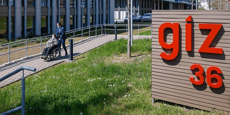 Helle Deertz im Freien vor einem Bürogebäude. Ihr Assistent schiebt ihren Rollstuhl über einen gepflasterten Weg. Im Vordergrund ein roter Schriftzug giz 36.