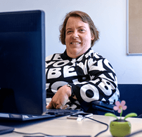 Helle Deertz sitzt am Schreibtisch. Sie trägt einen schwarz-weißen Pullover und lächelt in die Kamera. Im Vordergrund ein Computerbildschirm.