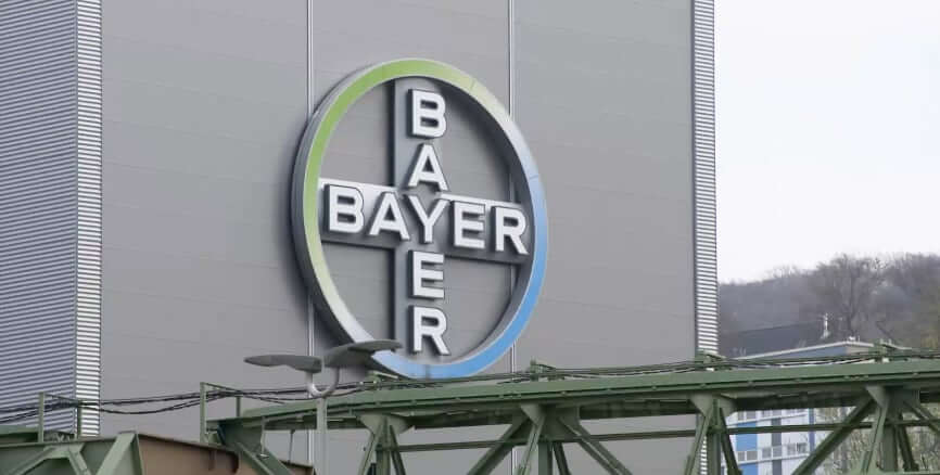Das Betriebsgebäude von Bayer mit den Bayer-Logo. Ein blau-grün-weißer Kreis. Innen als Kreuz zweimal der Name Bayer. Einmal von links nach rechts und einmal von oben nach unten geschrieben.