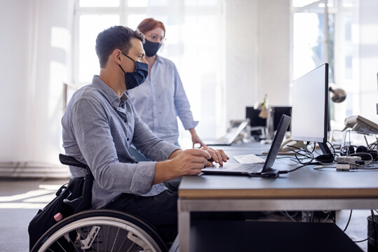 Mann mit Mundschutz im Rollstuhl sitzt an einem Schreibtisch. Neben ihm steht eine Frau mit Mundschutz. Beide schauen auf einen Computerbildschirm.