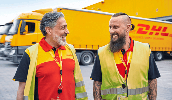 Zwei DHL Mitarbeiter lächeln sich an. Sie stehen vor einem DHL LKW.