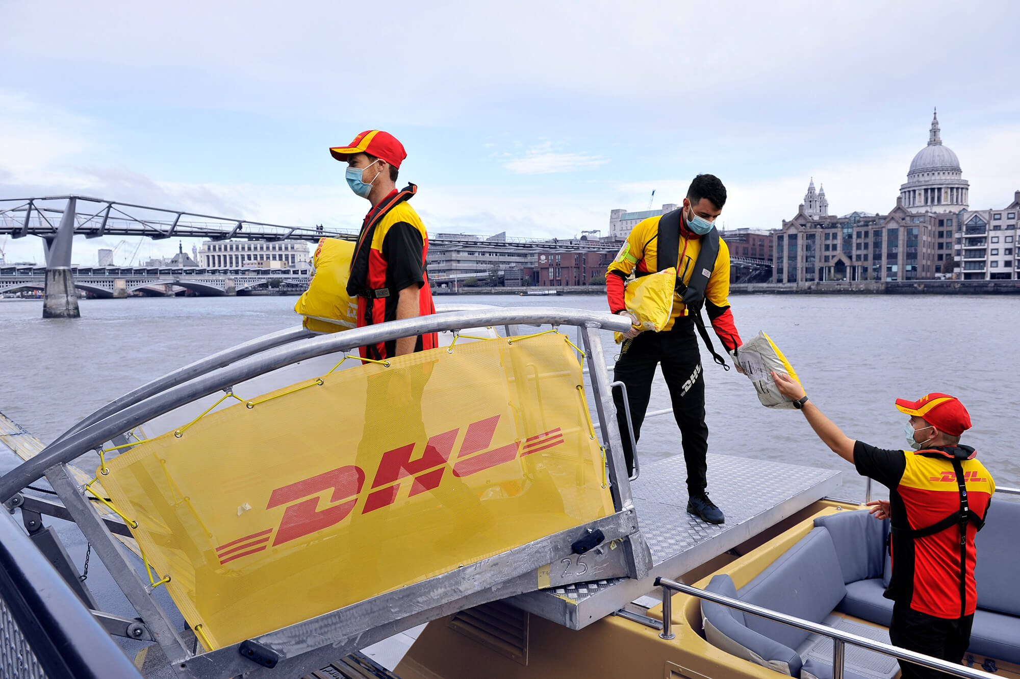 Drei DHL Mitarbeiter entladen ein kleines Schiff. Sie tragen DHL Arbeitskleidung in rot und gelb.
