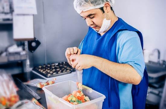 Ein junger Mann in blauem Arbeitskittel und mit Hygieneschutz. Er sortiert Fruchtgummi in einen Folienbeutel.