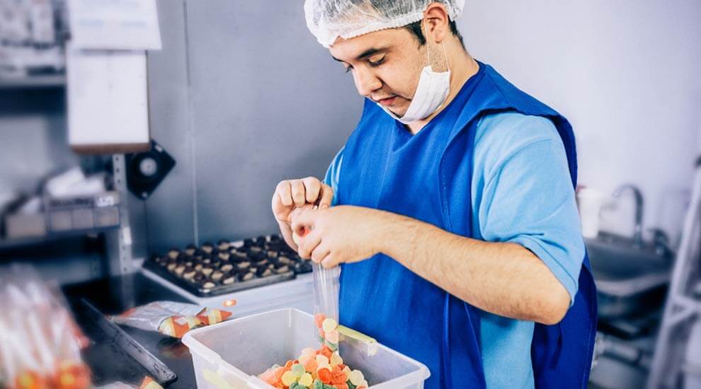 Ein junger Mann in blauem Arbeitskittel und mit Hygieneschutz. Er sortiert Fruchtgummi in einen Folienbeutel.