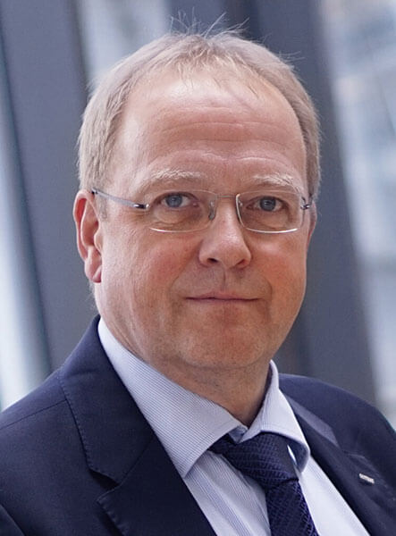 Portraitfoto von Mathias Hüsing. Er trägt eine Brille und einen dunkelblauen Anzug mit Hemd und Krawatte.  