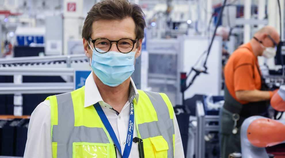 Christoph Beyer steht in Warnweste und mit Mundschutz in der Werkhalle bei Ford. Im Hintergrund sieht man Mitarbeiter am Fließband arbeiten.