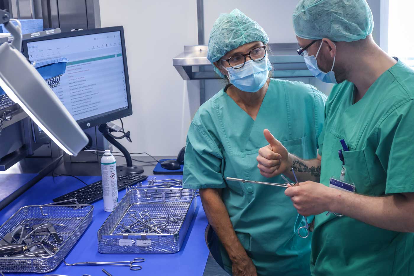 Zwei Mitarbeitende sind im Gespäch. Beide tragen Kittel, Haube und Maske. Einer der Mitarbeiter hat ein OP-Instrument in der Hand. Auf dem Arbeitsplatz vor ihnen liegen Körbe mit weiterem OP-Besteck. 