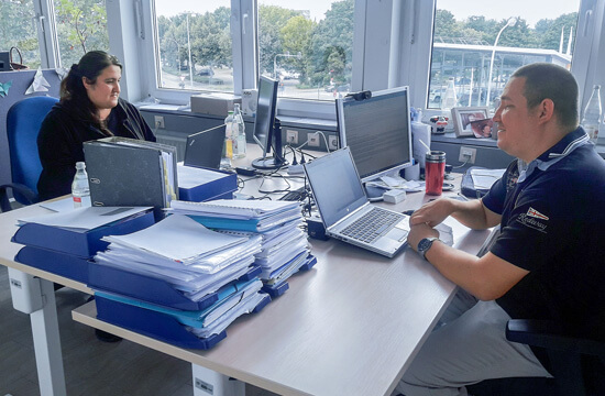 Eine Frau und ein Mann sitzen an zwei Schreibtischen gegenüber. Sie arbeiten am Laptop.