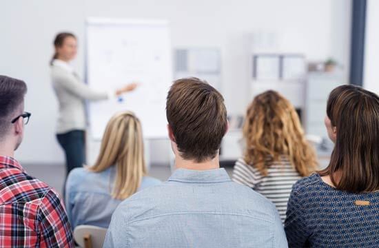 Ein Seminarraum. Sitzende Menschen von hinten. In Hintergrund eine Frau am Flipchart.