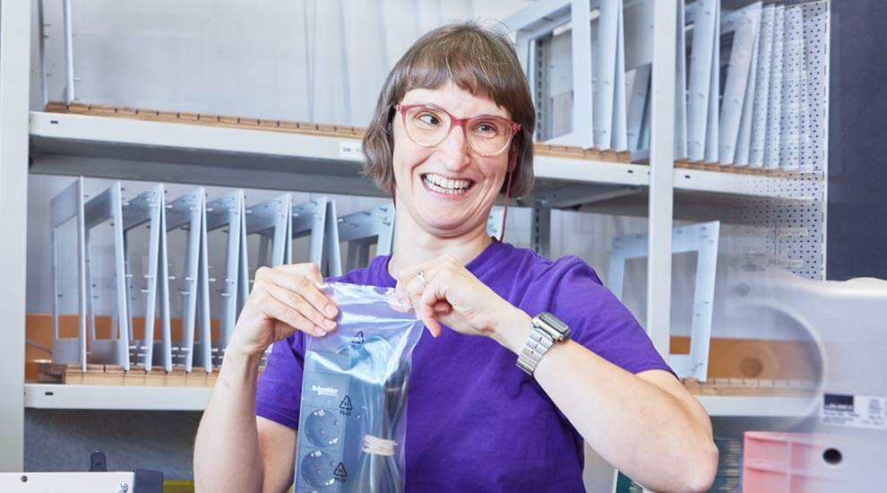 Eine Frau sitzt an einem Arbeitstisch. Sie hält eine eingeschweißte Steckerleiste vor dem Oberkörper.