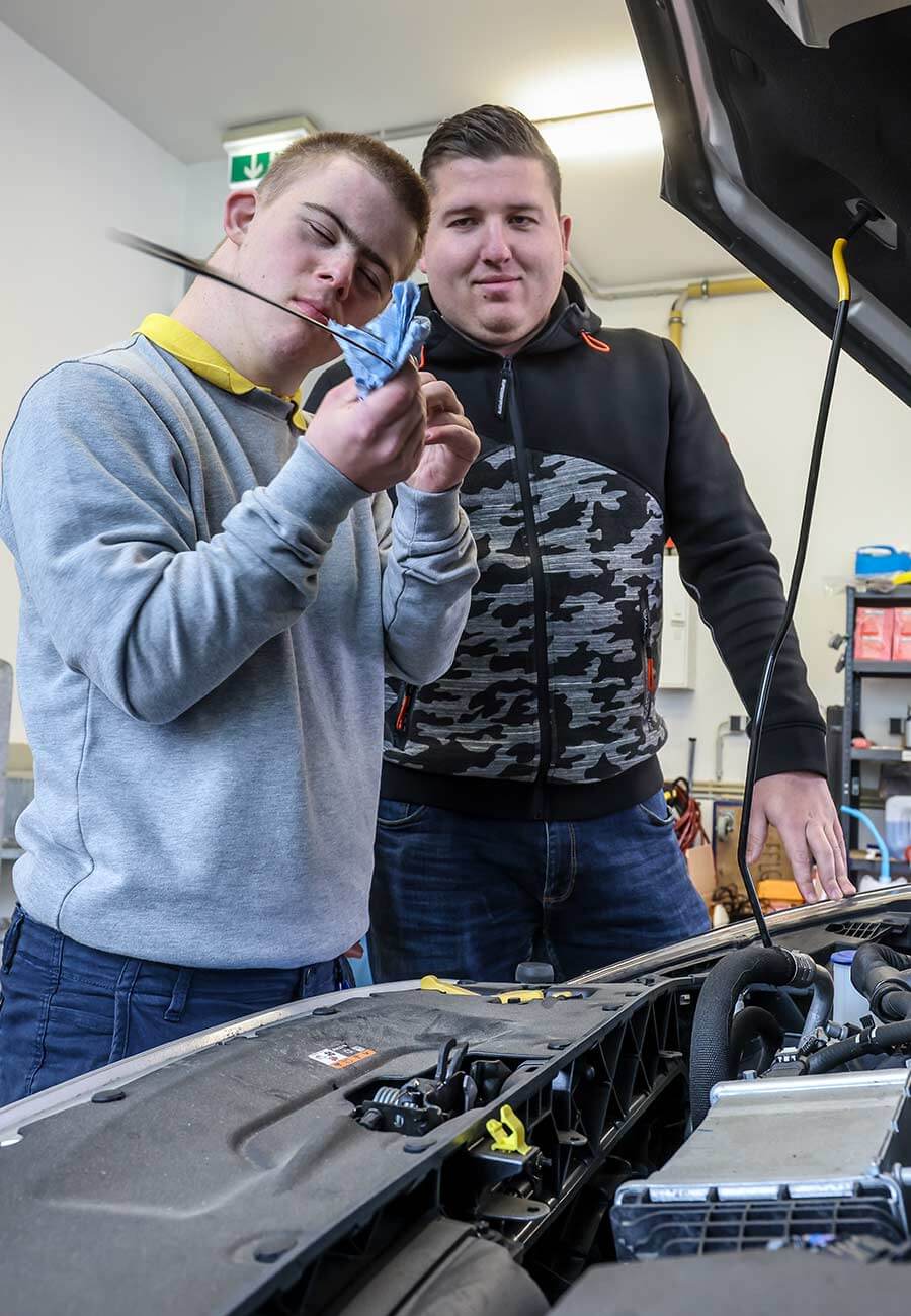 Eine Kfz-Werkstatt. Eine offene Motorhaube. Dominik überprüft den Ölmessstab. Neben ihm steht sein Kollege.