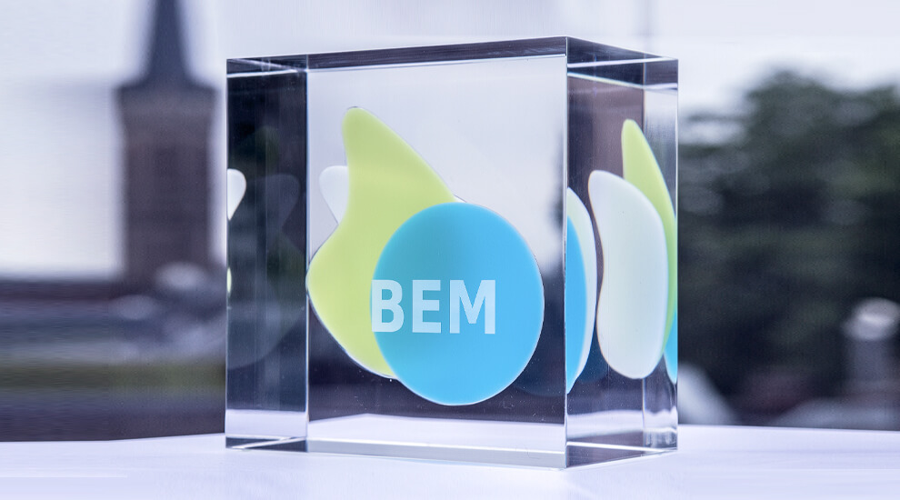 Durchsichtiger Quader mit blau-grün-weißem BEM-Logo auf einem Tisch im Freien.