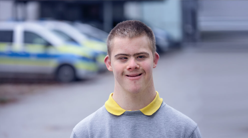 Portraitfoto von Dominik. Er lächelt. Im Hintergrund mehrere Polizeiautos.