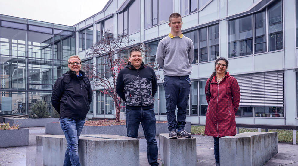 Gruppenbild mit vier Personen im Freien mit Dominik in der Mitte. Alle lächeln. Im Hintergrund ein Gebäude.