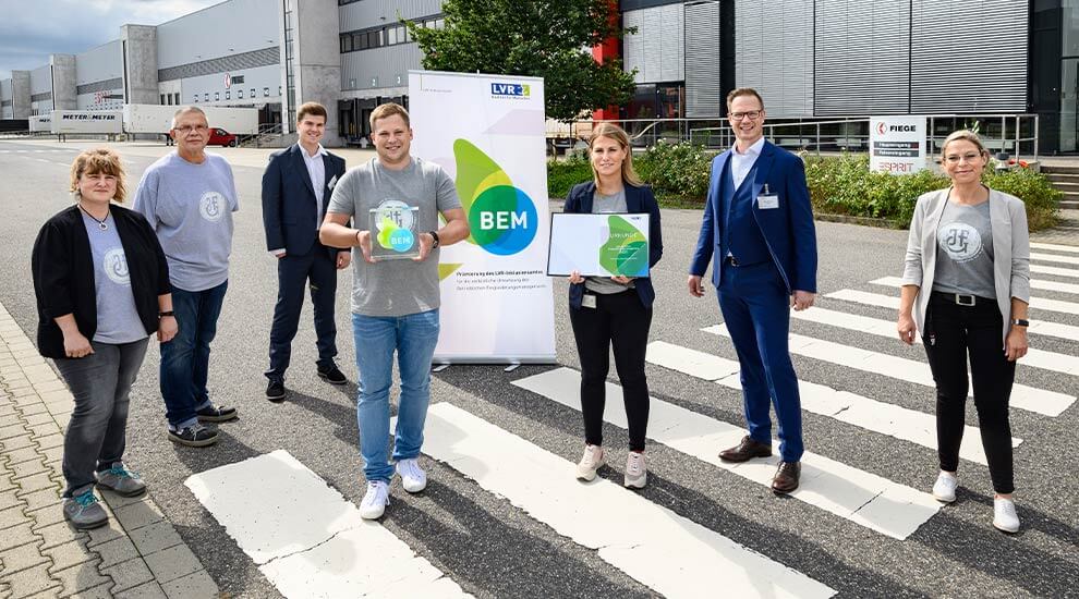 Gruppenbild im sieben Personen im Freien. Zwei Personen halten Auszeichnungen. Im Hintergrund ein gewerbliches Gebäude.