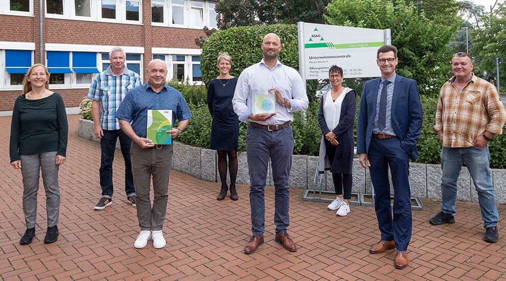 Gruppenbild mit acht Personen im Freien. Zwei Personen halten Auszeichnungen. Im Hintergrund ein Bürogebäude.