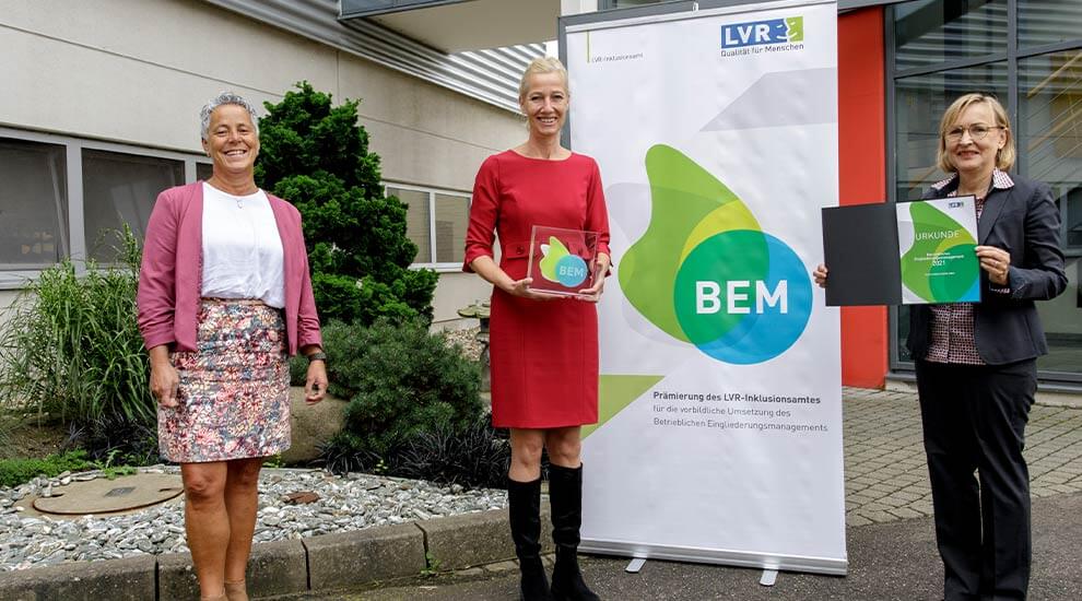 Gruppenbild mit drei Frauen vor dem Eingang eines Bürogebäudes. Zwei Frauen halten Auszeichnungen.