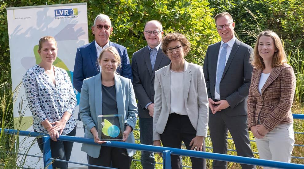 Gruppenbild mit sieben Personen im Freien. Eine Frau hält eine Auszeichnung.