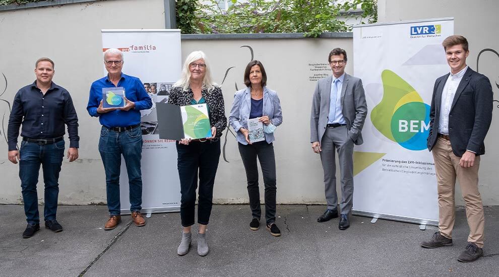 Gruppenbild mit sechs Personen. Sie stehen im Freien vor einer Mauer. Drei Personen halten Auszeichnungen. Rechts und links zwei Aufsteller mit Bannern.