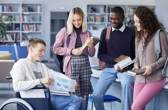 Eine Gruppe von vier jungen Menschen mit Büchern in einem Raum mit Bücherregalen. Ein Mann sitzt im Rollstuhl.