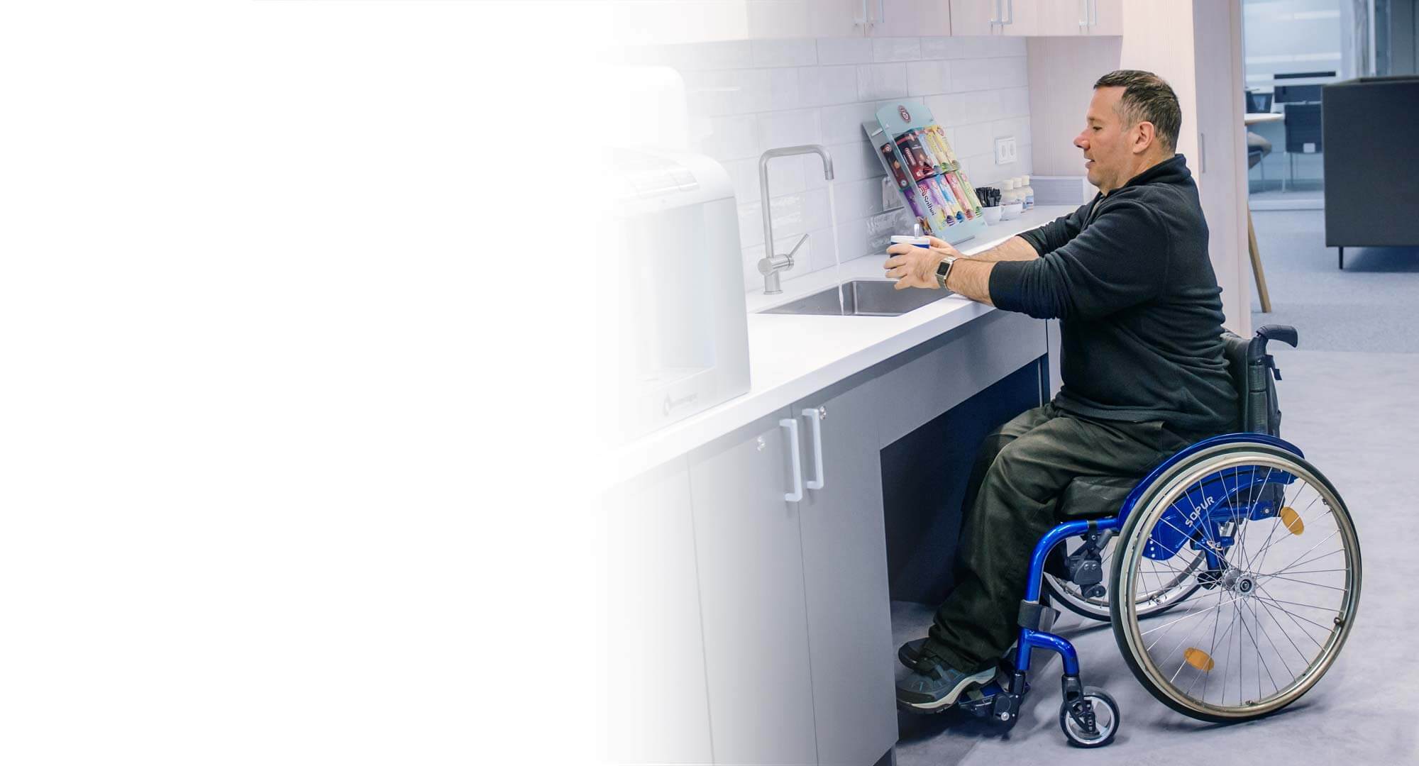 Mann in Rollstuhl hält eine Tasse unter einen Wasserhahn.