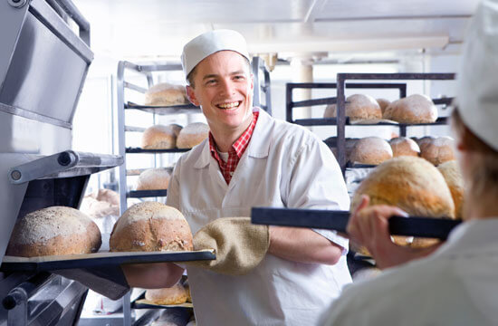 Junger Bäcker lächelt zu einem Kollegen. Er trägt Berufskleidung und holt Brote aus dem Ofen.