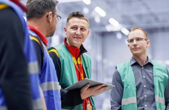 Vier DHL Mitarbeiter stehen in einem Lager. Sie tragen Warnwesten.