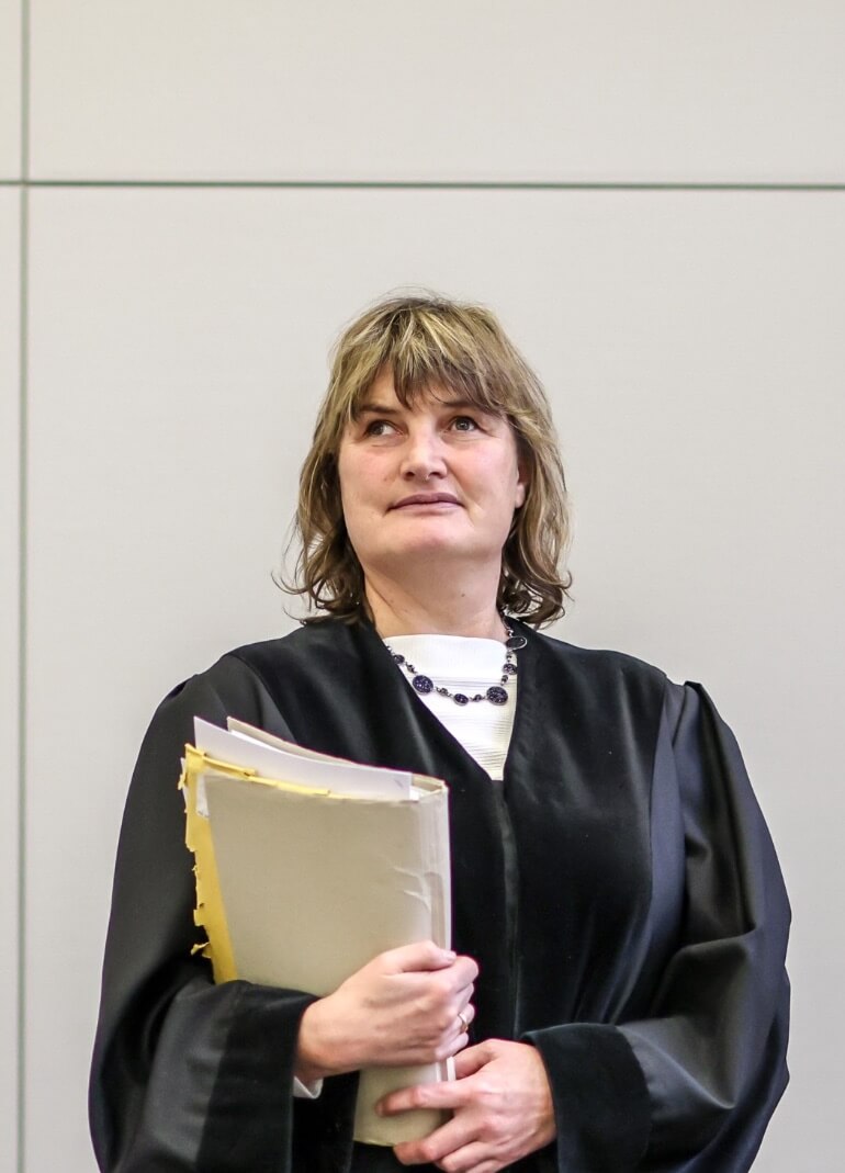 Richterin Dr. Bungart steht in schwarzer Robe mit Akten auf dem Arm in einem Gerichtssaal.