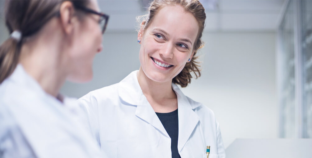 Zwei Frauen in weißen Kitteln lächeln sich zu und stehen in einem Labor.