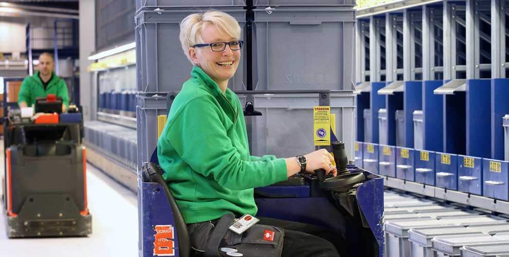 Blonde Frau mit grüner Jacke fährt Gabelstapler. Sie lächelt in die Kamera.