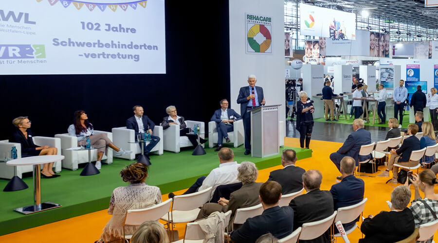 Das Foto zeigt eine seitliche Sicht auf die Bühne in einer Messehalle. Mehrere Personen sitzen auf dem Podium vor einer Leinwand mit dem eingeblendeten Titel „102 Jahre Schwerbehindertenvertretung“. Rechts davon steht ein Mann am Rednerpult. Im Vordergrund ist das Publikum zu sehen.