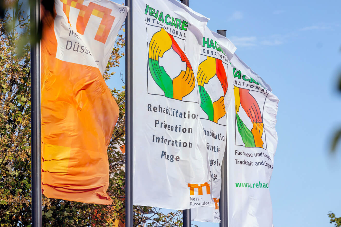 Das Foto zeigt vier wehende Fahnen: Die erste zeigt das Logo der Messe Düsseldorf auf orangenem Grund. Die drei weißen Fahnen dahinter bewerben die Messe „Rehacare“.