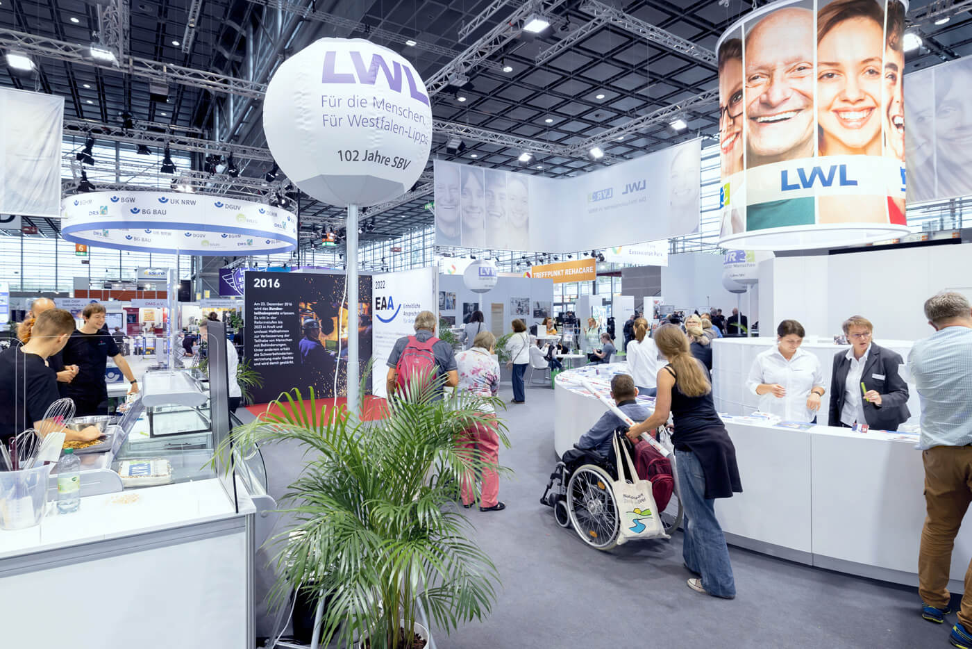 Das Foto zeigt den weitläufigen Messestand von LWL und LVR. Im Gang ist eine junge Frau zu sehen, die eine Person im Rollstuhl schiebt.