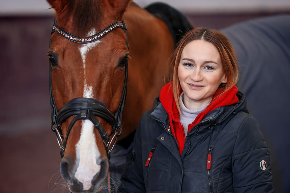 Das Foto zeigt eine junge Frau, die ein braunes Reitpferd mit Strass geschmücktem Zaumzeug am Zügel hält.