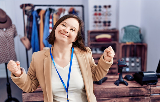 Eine Frau mit Down-Syndrom ballt lächelnd die Fäuste; im Hintergrund ist ein Modeatelier zu sehen