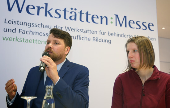 Vor einem Plakat mit der Aufschrift "Werkstättenmesse" stehen ein Mann und eine Frau; der Mann spricht in ein Mikrofon.