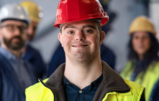 Ein Mann mit Down-Syndrom in Arbeitskleidung und mit Schutzhelm steht vor einer Gruppe weiterer Arbeiter 