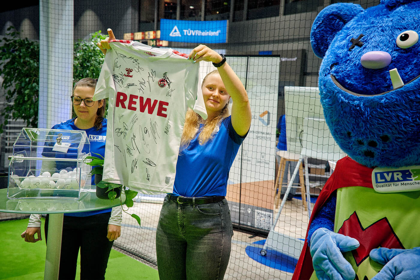 Zwei junge Frauen stehen an einem Messestand vor einer gläsernen Lostrommel. Die Frau rechts hält ein signiertes Trikot des 1. FC Köln hoch. 