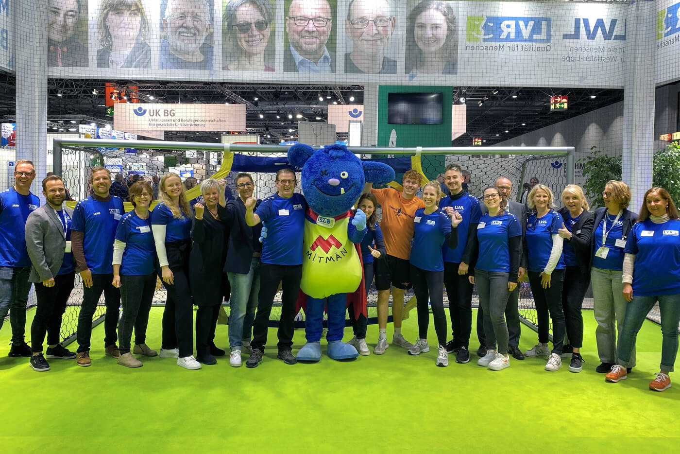 Gruppenbild des LVR/LWL-Messeteams vor dem am Messestand aufgebauten Fußballtor. In der Mitte steht das LVR-Maskottchen Mitmän.