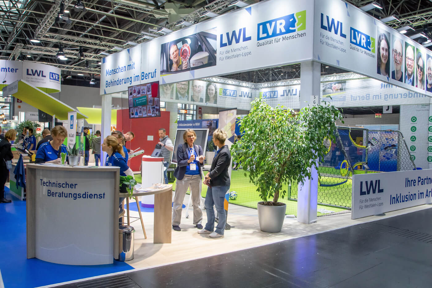 Der LVR/LWL-Messestand, vorne ein Tresen des Technischen Beratungsdienstes im Hintergrund das Fußballfeld mit Fußballtoren. Einige Mitarbeiter sind im Gespräch mit Messebesuchern