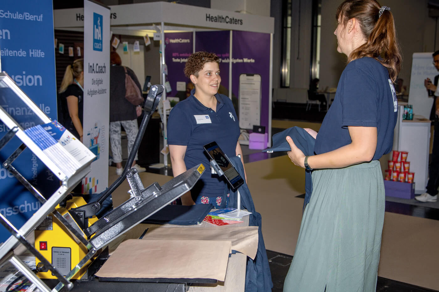 Zwei Frauen stehen an einem Messestand, vor ihnen ist eine Druckmaschine zum Bedrucken von dunkelblauen Stofftaschen zu sehen.
