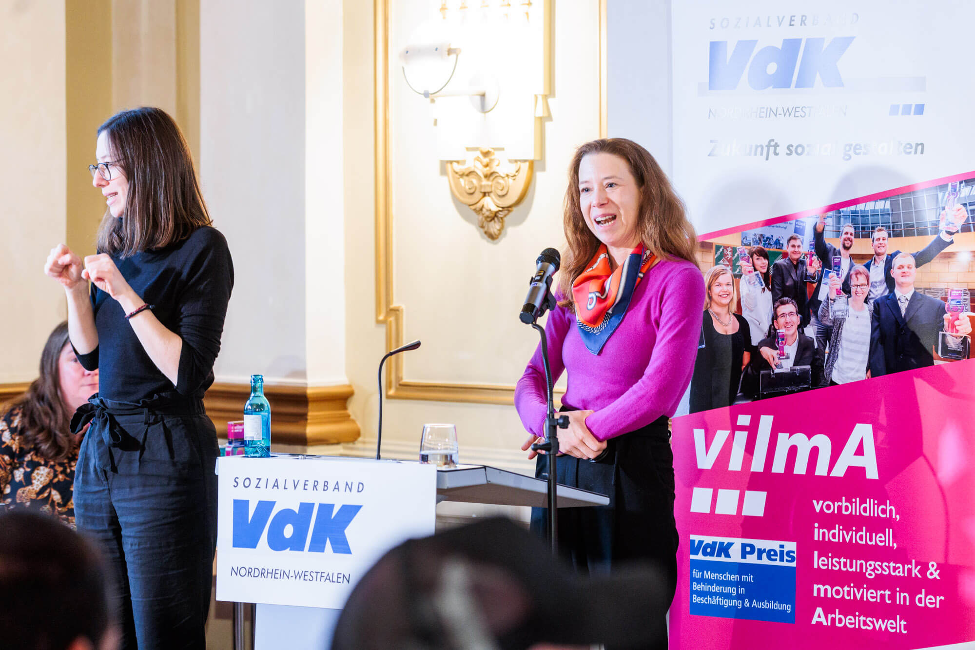 Melanie Glücks vom im LVR-Inklusionsamt steht bei der VdK- Preisverleihung auf einer Bühne vor einem Mikrophon, neben ihr steht noch eine Gebrädendolmetscherin.