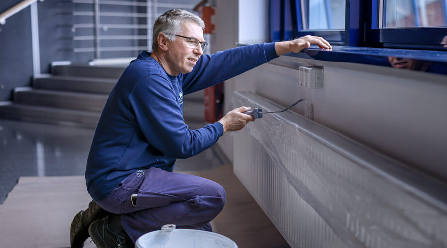 Maler Stefan Schulz kniet auf einem Knie auf dem abgedeckten Boden und streicht eine Wand unter einem Fensterbrett. 