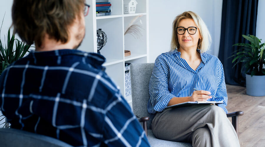 BEM | Foto: shutterstock/Okrasiuk Eine Frau sitzt in einem Büroraum in einem Sessel, auf ihrem Schoß liegt ein Klemmbrett. Sie schaut ihren Gesprächspartner freundlich an. Der Gesprächspartner ist nur schräg von hinten am linken Bildrand zu sehen. Im Hintergrund steht ein offenes weißes Regal.