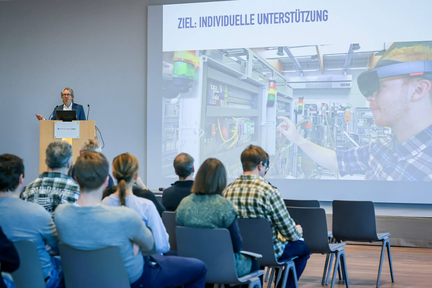 Prof. Dr. Carsten Röcker steht hinter einem Rednerpult auf einer Bühne. Vor der Bühne sitzen die Zuhörer auf Stühlen. 