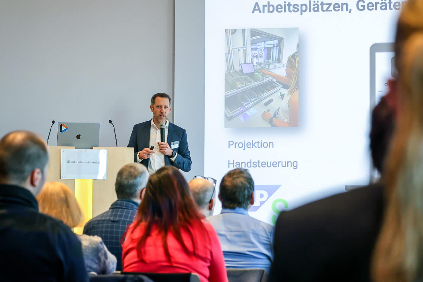 Eckhard Bickler bei seinem Vortrag auf einer Bühne. Vor ihm das Publikum zu sehen, hinter ihm ist eine Präsentation an eine große Leinwand projeziert.rt.