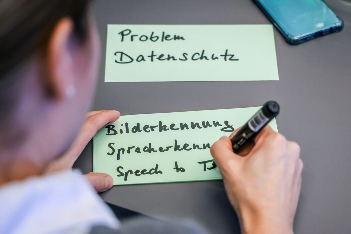Nahaufnahme über die Schulter einer Workshopteilnehmerin, die gerade mit einem Permanentmarker etwas auf einen grünen Zettel schreibt.