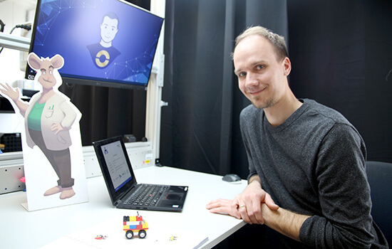 Der Projektkoordinator sitzt am Arbeitstisch. Darauf ein Laptop, ein Aufsteller der Comic-Maus Gregor im Anzug und das H-Auto aus Legosteinen.