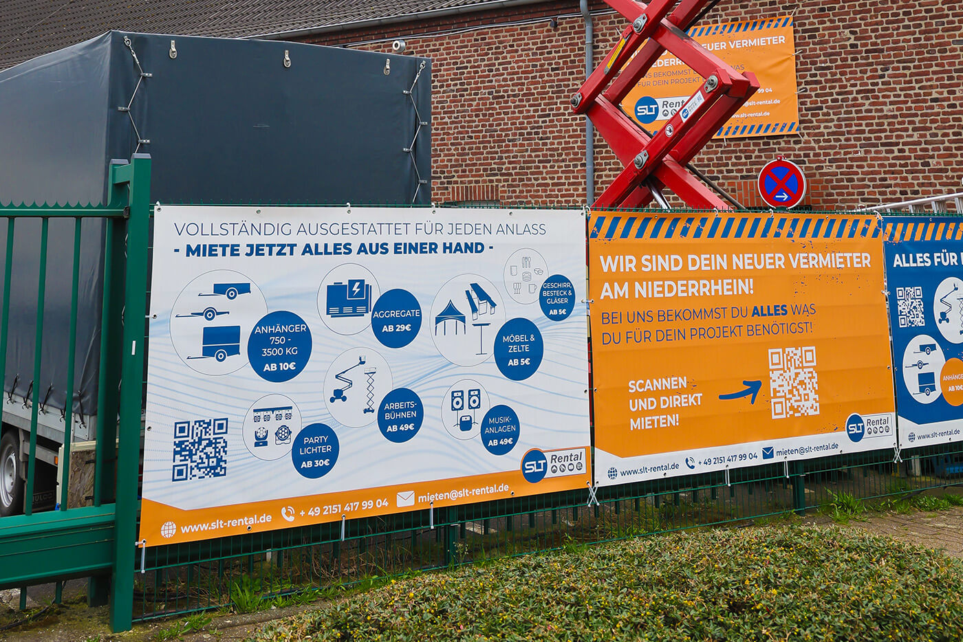 Ein weißes und ein orangenes Banner an einem Zaun schildern die Dienstleistungen der Firma SLT.